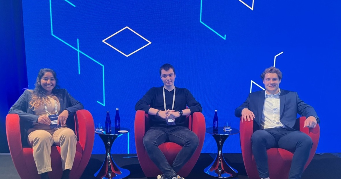 Three students with their conference badges, each in a red lounge chair in front of a graphic blue wall. Pictured are Mira Manchanda, Filip Matic, and Clayton Burton. 
