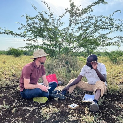 Evan Griffith, wearing a sunhat, sits with a colleague under the shade of a small tree in Turkana, Kenya.