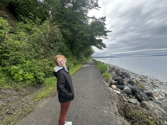 MAddie stands on a paved path with trees on one side and water on the other